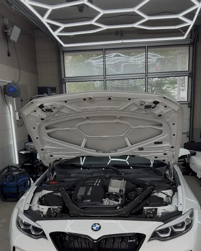 Weißer BMW M2 mit geöffneter Motorhaube in der Halle unter dem Wabenlicht